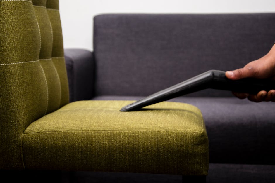 A close-up image of a person's hand holding a vacuum hose attachment, performing surface cleaning on a beige fabric car seat with a patterned, slightly textured surface. The seat appears slightly dirt