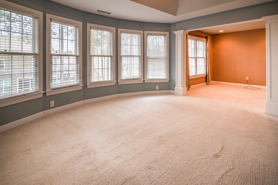 A person using a vacuum cleaner with a yellow body and black hose to perform surface cleaning and deep cleaning of an ornate, patterned area rug in a home setting. The individual is wearing casual clo