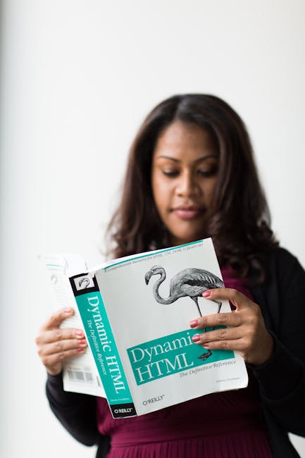 A woman with long, dark hair is reading a book titled 'Dynamic HTML' by O'Reilly, holding the book with both hands. The book features a cover with a detailed illustration of a flamingo. She is standin